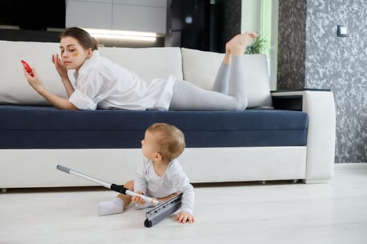 Baby boy play with mop, while mother surfing in your smartphone lying on couch.