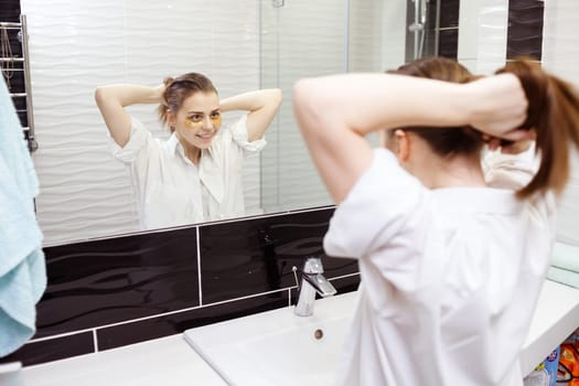 Young woman with hydrogel patches under eyes makes ponytail looking into the mirror in bathroom.