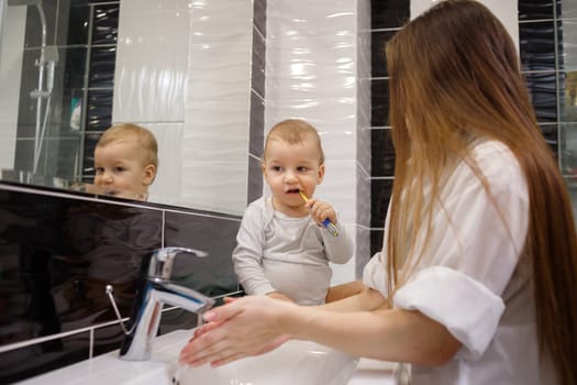 Child brushing teeth sitting on sink near mirror in bathroom. His mother monitoring action.