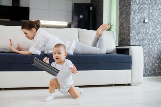 Baby boy play with mop, while mother surfing in your smartphone lying on couch
