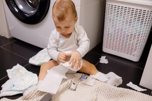 the child sits on a white toilet. in his hands is toilet paper. there are a lot of rolls on the floor. close-up