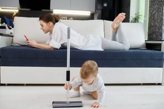 Baby boy play with mop, while mother surfing in your smartphone lying on couch.