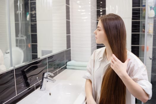 Young woman combing her long hair in bathroom after getting up in the morning. Young attractive happy woman is brushing her brown hair enjoying them in the mirror and admiring herself