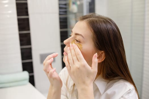 Caucasian woman applying hydrogel patches on under eye area, looking at mirror. Happy young lady enjoying morning anti age anti wrinkle routine in bathroom.