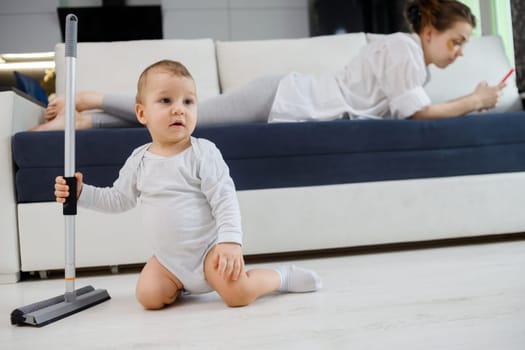 Baby boy play with mop, while mother surfing in your smartphone lying on couch.