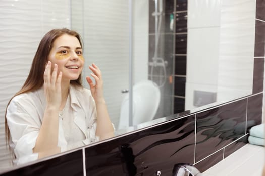 Caucasian woman applying hydrogel patches on under eye area, looking at mirror. Happy young lady enjoying morning anti age anti wrinkle routine in bathroom