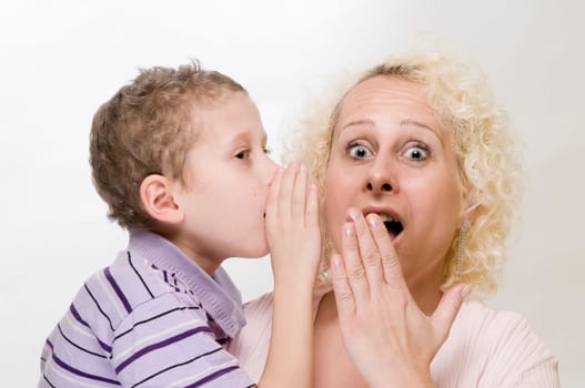 Head-and-shoulder view of a 7-year-old boy whispering into his blonde mother's ear with his hand held in front of a white background  Mother looks into the camera in shock