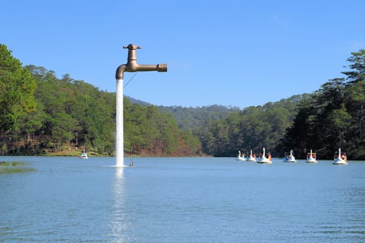An oversized faucet in the middle of the lake appears to be floating above the water of a lake with swan boats in Dalat, Vietnam