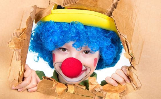 young child dressed as a clown with wig and fake nose has fun watching from the open cardboard
