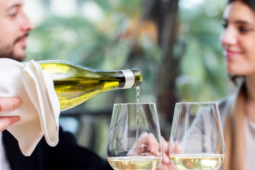 Close up of waiter pouring white wine in glass with couple in background.