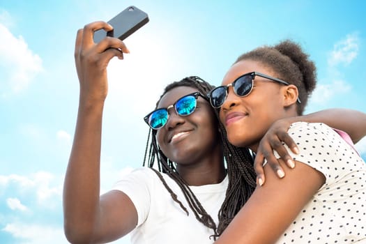 Close up portrait of two diverse african teen girls wearing sun glasses taking self portrait with phone against blue sky.