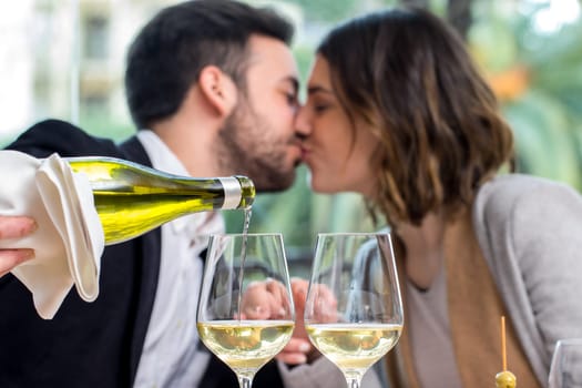 Close up of hand pouring white wine in glass with romantic out of focus couple in background.