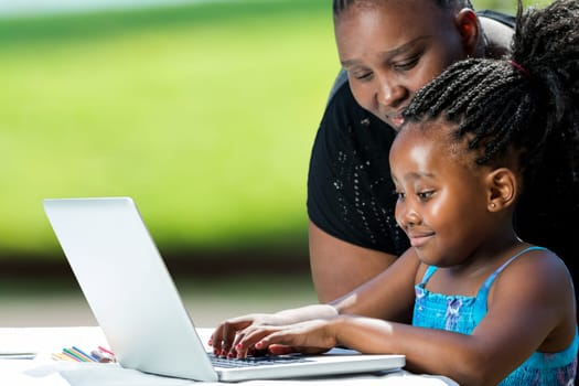 Close up portrait of african mother supervising school work of little daughter on laptop. Cute girl typing on laptop against green background outdoors.