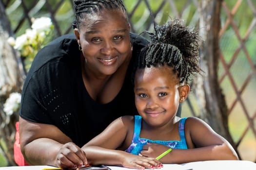 Close up portrait of african mother and little daughter with braided hairstyle in garden.Girl drawing on paper with color pencils outdoors.