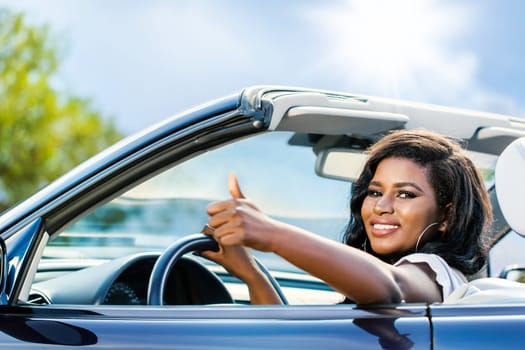 Close up portrait of attractive black female driver doing thumbs up.Young girl sitting in convertible.