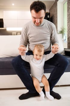 Young father assisting walking his infant son teaching to walk at home. baby boy learns to walk with daddy's support