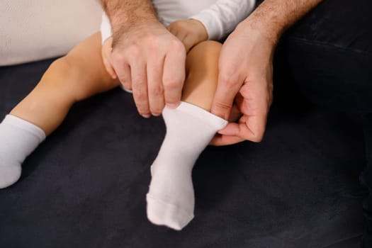 Young father is putting socks on his baby son, sitting on sofa.