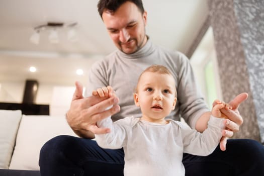 Young father assisting walking his infant son teaching to walk at home. Daddy helps son attempting to walk.