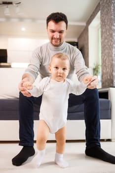 Young father assisting walking his infant son teaching to walk at home. Daddy helps son attempting to walk