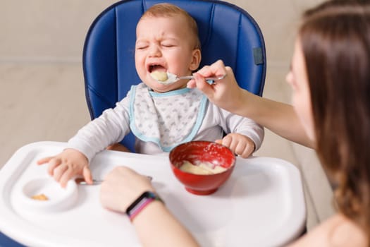 Mother tries to feed with spoon naughty infant kid refusing eating.