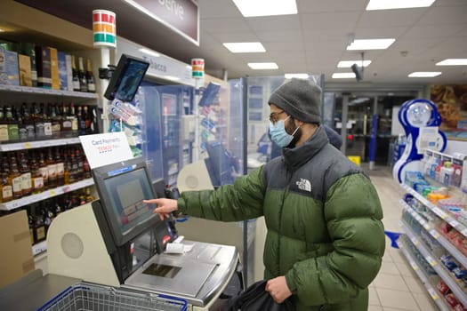 Sheffield, United Kingdom, 14th January, 2021: Young man wearing a face mask and winter clothes pays for groceries at a Tesco express self checkout