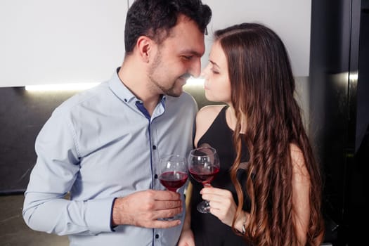Close up couple of lovers holding glasses with red wine, touching foreheads, enjoying romantic date, standing in kitchen, celebrating relocation or anniversary or valentines day.