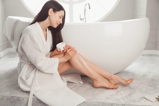Brunette woman sitting at bathtub and applying cream on legs.