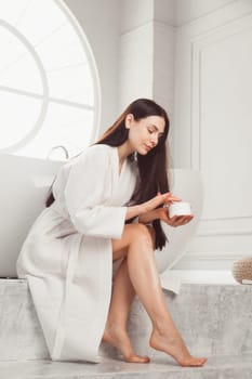 Young brunette woman applying body scrub or cream on skin sitting in luxury bathroom.