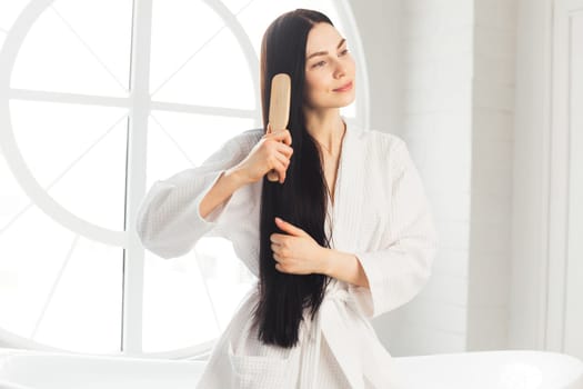 Young brunette woman combing her healthy and shiny hair sitting on bathtub weared in white bathrobe.