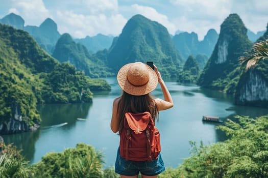 A female traveler in a hat and a T-shirt with a backpack on her back takes a photo of an amazing landscape with rocks, sea and blue sky on her smartphone during her vacation.