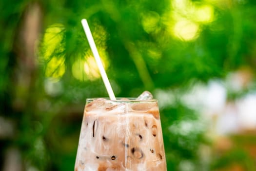 Close up summer refreshment drinks. Chilled iced chocolate cocoa. Chocolate powder and ice. In glasses, with tubes for drinking. Green leaves nature background. Close up of a glass of Iced Cocoa cool drink served on table. Iced chocolate drink in classic glass on green nature of summer background. Summer drink