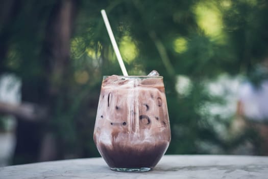 Close up summer refreshment drinks. Chilled iced chocolate cocoa. Chocolate powder and ice. In glasses, with tubes for drinking. Green leaves nature background. Close up of a glass of Iced Cocoa cool drink served on table. Iced chocolate drink in classic glass on green nature of summer background. Summer drink