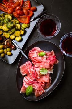 Prosciutto with different appetizers with glass of red wine on black background. Over roasted vegetables, healthy snack