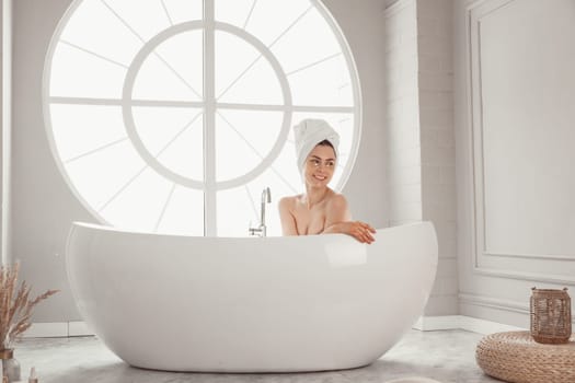 Young woman relaxing lying in bathtube in luxury bathroom with round large window.
