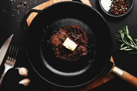Cooked ribeye steak on cast-iron pan and board with pepper, rosemary, salt, garlic, knife, fork, butter on black background