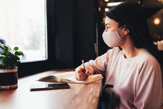 Girl in face mask, watching in window, writing in notebook. Blurred background. High quality photo