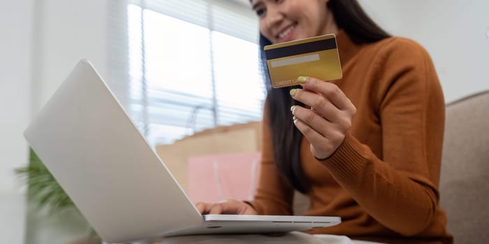 A delighted woman holds her credit card while using a laptop for online shopping, surrounded by colorful shopping bags.
