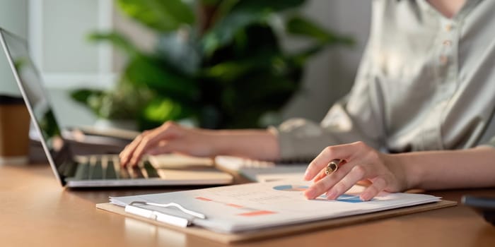 A financial analyst examines charts on a clipboard and works on a laptop, focusing on data analysis and budgeting.