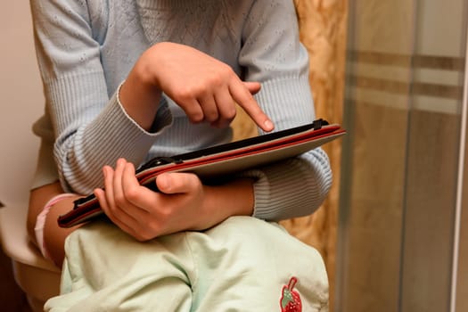 The child sits on the toilet and plays on the tablet