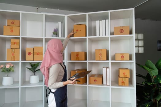 A Muslim woman carefully organizes packages on a shelf in her home office, preparing for her online sales business.