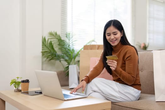 A joyful woman uses her laptop for online shopping, holding a credit card and surrounded by shopping bags, expressing excitement.