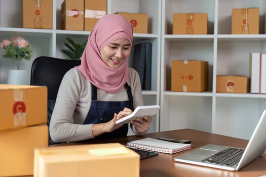 A happy Muslim woman in a pink hijab checks her online business inventory while preparing packages, reflecting her commitment to entrepreneurship.