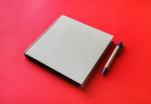 Photo of closed blank square book and pen on red paper background. Template for placing your design.