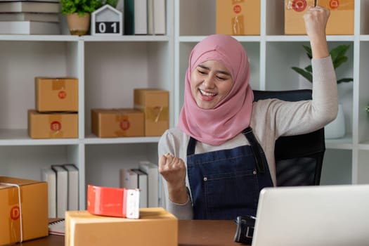 A joyful Muslim woman celebrates a milestone in her online business, expressing excitement in her organized home office.