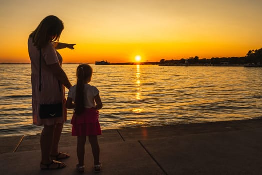 Mother showing her daughter a beautiful sunset at the beach while holding hands. Silhouette of a marina with boats at Adriatic Sea in Zaton, Croatia