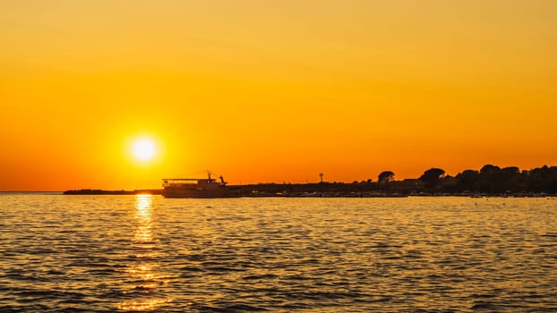 Amazing sunset with defined Sun and reflection on water. Silhouettes of boats and cruise ship moored at marina on Adriatic Sea, Zaton, Croatia