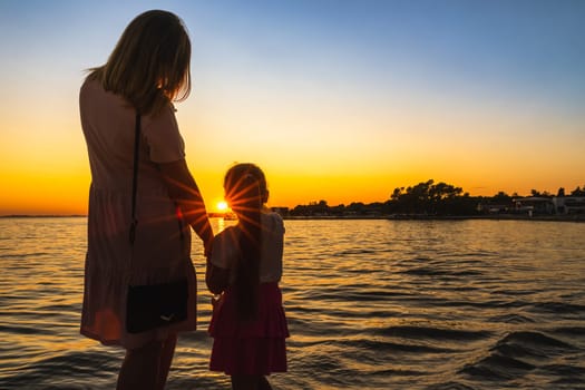 Mother and daughter holding hands, admiring beautiful sunset at Adriatic Sea beach. Silhouette of a family which trough shines Sunstar, Zaton, Croatia