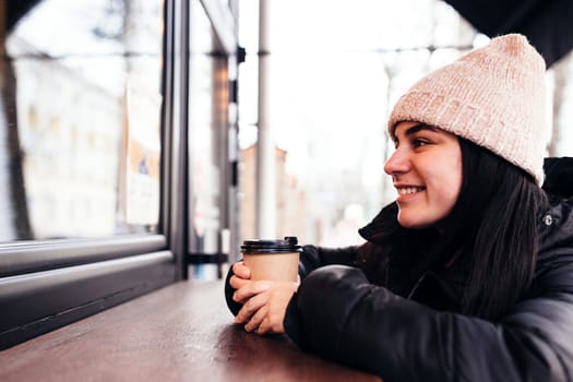 Girl smiling, holds paper cup of coffee in hand near cafe. Blurred background. High quality photo