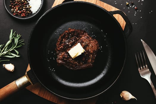 Cooked ribeye steak on cast-iron pan and board with pepper, rosemary, salt, garlic, knife, fork, butter on black background