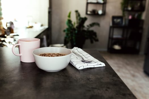Pink coffee cup, bowl with chopped tropical fruits kiwi and banana, blueberries, spoon spoon on towel on bar counter in stylish loft kitchen. Blurred background. High quality photo
